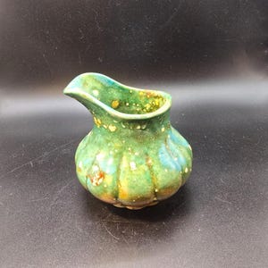 Puede incluir: Una pequeña jarra de cerámica hecha a mano con un cuerpo bulboso y un pico. La jarra está esmaltada en tonos verdes, azules y amarillos, con acentos moteados. El diseño es orgánico y rústico, lo que sugiere una estética artesanal.