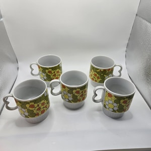 May include: Five white ceramic mugs with a floral pattern in yellow, orange, and green. The mugs have a rounded base and a unique handle design. The floral pattern is repeated around the body of each mug.
