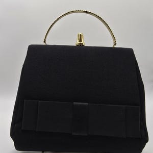 以下が含まれることがあります： ゴールドトーンのハンドルと留め金が付いた黒いハンドバッグ。長方形のバッグは前面に大きなリボンが付いています。ハンドルはアーチ型で、小さなキラキラとしたアクセントで装飾されています。バッグは無地の白い背景に置かれています。
