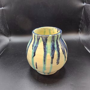 Puede incluir: Un pequeño jarrón de cerámica redondo con una abertura ancha. El jarrón tiene una base de color crema con vetas verticales de pintura azul y verde azulado. El interior del jarrón es de color amarillo pálido.