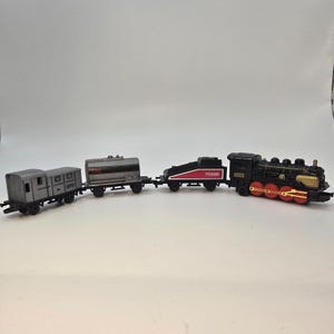 May include: A toy train set featuring a black and red locomotive with gold accents, pulling three cars. The cars include a gray boxcar, a silver tanker car labeled "GAS", and a black car with red detailing and the number "703698".