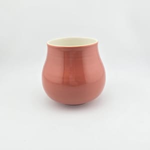 May include: A rounded ceramic cup with a smooth, glossy finish in a muted dusty rose color. The vessel features a wide, bulbous body that tapers slightly toward the top rim, revealing a clean, off-white interior surface against the exterior pink.