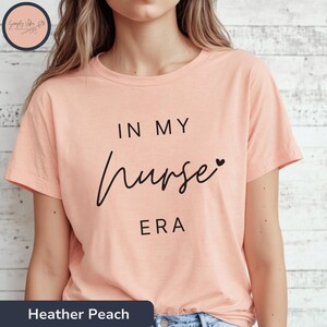 Könnte beinhalten: Ein pfirsichfarbenes T-Shirt mit dem Text "In My Nurse Era" in schwarzer Schrift mit einem kleinen Herzsymbol.