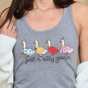 K&ouml;nnte beinhalten: Graues Tanktop mit einer skurrilen Illustration von vier G&auml;nsen in verschiedenen Outfits, darunter ein Blumenkleid, ein gelber Mantel, ein Erdbeerkost&uuml;m und ein rosa Rock. Der Text "just a silly goose" steht darunter.