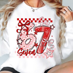 May include: White sweatshirt with a large graphic featuring the numbers "67" in red and pink, with the word "Six" above and "Seven" below. The design includes hearts, stars, and bows, with a checkered border.