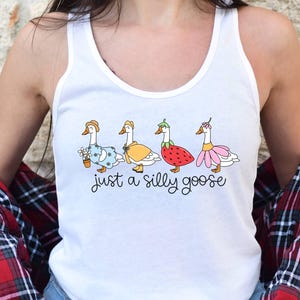 May include: White tank top with a whimsical illustration of four geese, each adorned with a different outfit, including a floral dress, a yellow dress, a strawberry dress, and a pink flower dress. The text "just a silly goose" is written below the geese.