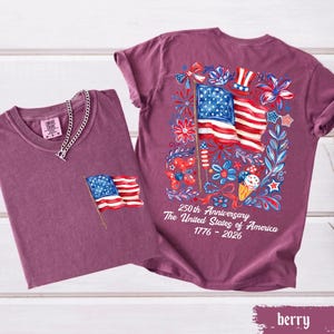 May include: Two berry-colored t-shirts. One has a small American flag on the pocket and a silver chain necklace. The other shirt has a large American flag graphic on the back with the text "250th Anniversary The United States of America 1776-2026".