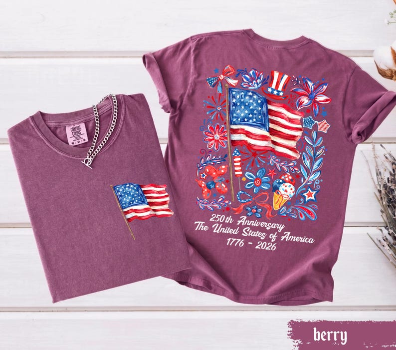 May include: Two berry-colored t-shirts. One has a small American flag on the pocket and a silver chain necklace. The other shirt has a large American flag graphic on the back with the text "250th Anniversary The United States of America 1776-2026".