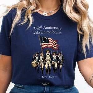 May include: Navy blue t-shirt commemorating the 250th Anniversary of the United States. The design features a waving American flag and a line of soldiers. The text "250th Anniversary of the United States" and the years "1776-2026" are also printed on the shirt.