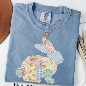 May include: A blue jean colored t-shirt with a patchwork bunny design. The bunny is made of floral and gingham fabrics in pink, yellow, and blue. A gold necklace with a small pendant is draped across the shirt. The shirt has a Comfort Colors tag.