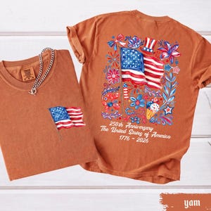 May include: Two rust-colored t-shirts. One has a small American flag on the pocket, the other has a large flag design with floral accents and text that reads "250th Anniversary The United States of America 1776-2026."