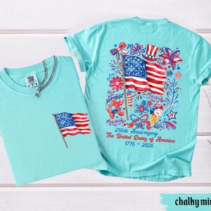 May include: Two chalky mint-colored t-shirts. One shirt has a small American flag graphic on the pocket. The other shirt has a large graphic of the American flag, flowers, and text that reads "250th Anniversary The United States of America 1776-2026."