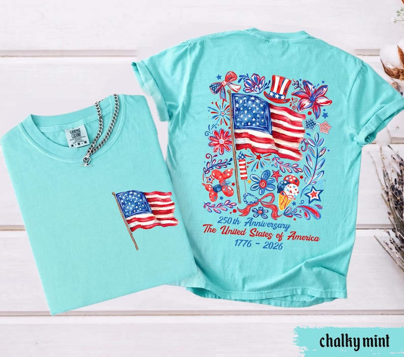 May include: Two chalky mint-colored t-shirts. One shirt has a small American flag graphic on the pocket. The other shirt has a large graphic of the American flag, flowers, and text that reads "250th Anniversary The United States of America 1776-2026."