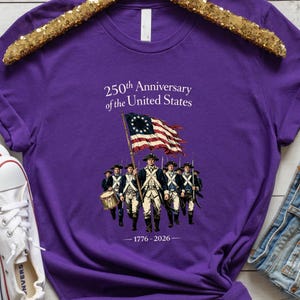 May include: Purple t-shirt featuring an illustration of soldiers carrying a flag with stars and stripes. The text reads "250th Anniversary of the United States" and "1776-2026". The shirt is displayed on a gold sequined hanger.