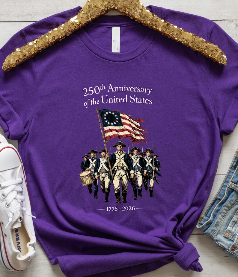 May include: Purple t-shirt featuring an illustration of soldiers carrying a flag with stars and stripes. The text reads "250th Anniversary of the United States" and "1776-2026". The shirt is displayed on a gold sequined hanger.