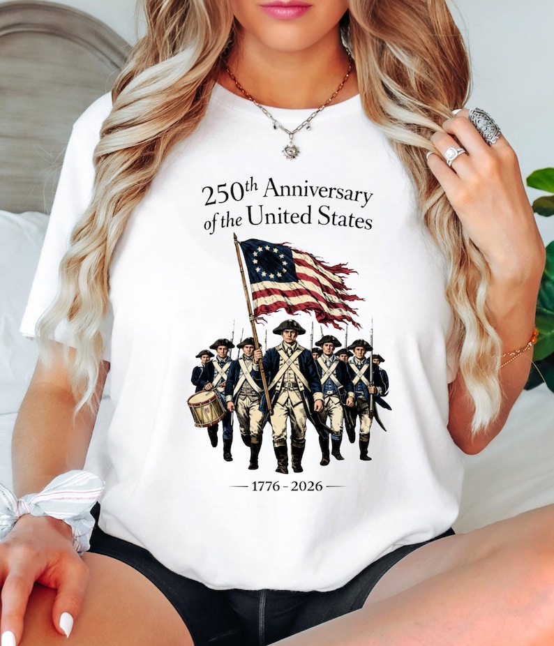 May include: White t-shirt featuring a graphic commemorating the 250th Anniversary of the United States. The design includes an illustration of soldiers marching with a flag and the text "250th Anniversary of the United States - 1776-2026."