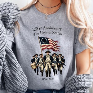 May include: A gray t-shirt with the text "250th Anniversary of the United States" and an illustration of soldiers carrying a flag. The flag is red and white striped with a circle of stars. The years 1776-2026 are printed below the illustration.