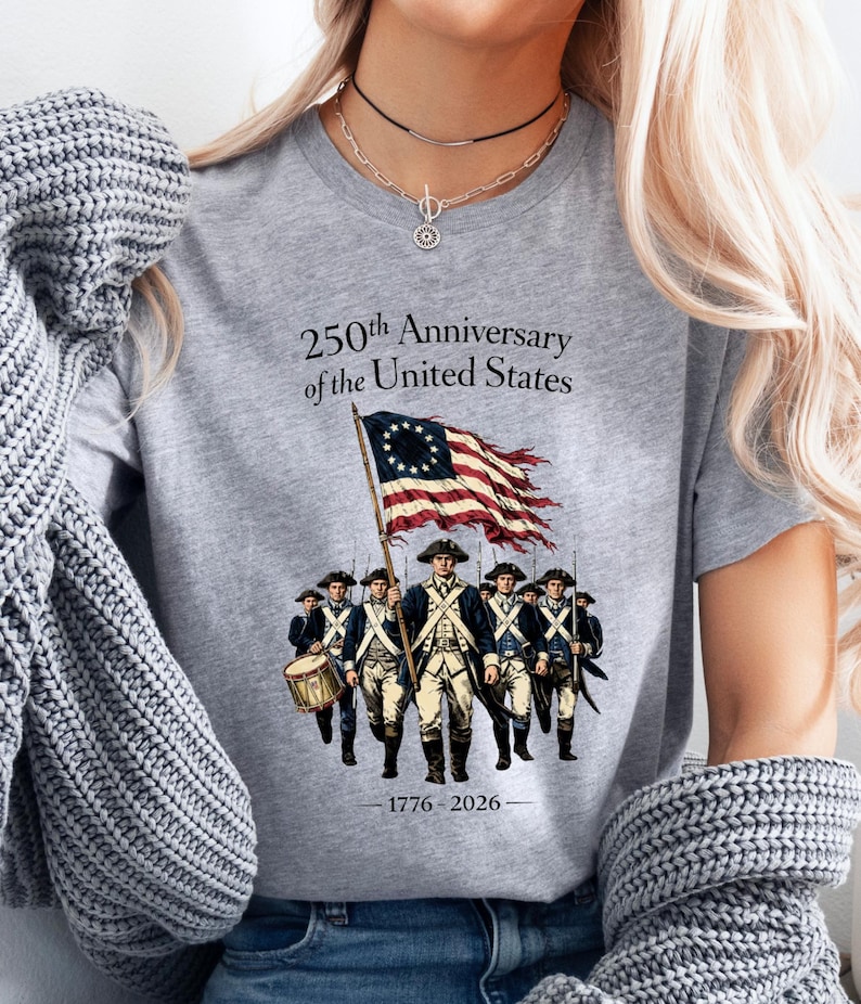 May include: A gray t-shirt with the text "250th Anniversary of the United States" and an illustration of soldiers carrying a flag. The flag is red and white striped with a circle of stars. The years 1776-2026 are printed below the illustration.