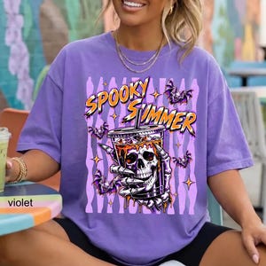 May include: A violet t-shirt featuring a graphic print of a skeletal hand holding a drink cup with a skull face. The text "SPOOKY SUMMER" is printed in orange above the cup, surrounded by small purple bats and star shapes against a vertical striped background.
