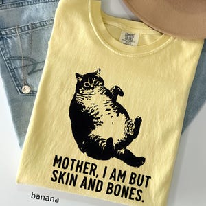 May include: Banana yellow t-shirt with a black graphic of a cat and the text "MOTHER, I AM BUT SKIN AND BONES." The shirt is folded and rests on a pair of blue jeans. A tan hat is visible in the background.
