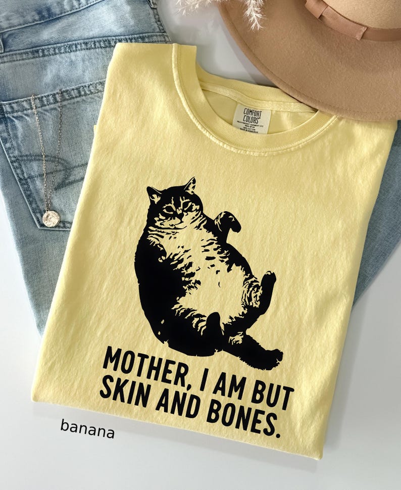 May include: Banana yellow t-shirt with a black graphic of a cat and the text "MOTHER, I AM BUT SKIN AND BONES." The shirt is folded and rests on a pair of blue jeans. A tan hat is visible in the background.