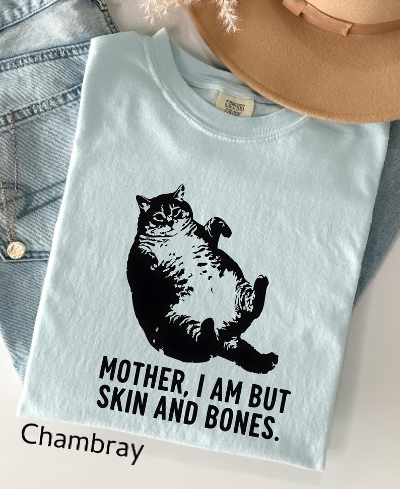 May include: Light blue t-shirt with a black graphic of a cat lying on its back. The text "MOTHER, I AM BUT SKIN AND BONES" is printed below the cat. The word "Chambray" is printed at the bottom left.