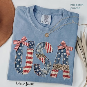 May include: A light blue Comfort Colors t-shirt with the word "USA" appliqued on the front. The letters are made of denim, red and white striped fabric, and leopard print fabric. Each letter is decorated with a red and white striped bow. The text "blue jean" is at the bottom.