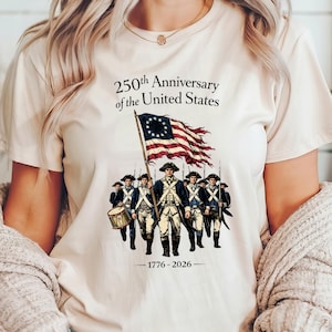 May include: Cream-colored t-shirt featuring a graphic commemorating the 250th Anniversary of the United States. The design includes an illustration of soldiers, a flag, and the years 1776-2026.