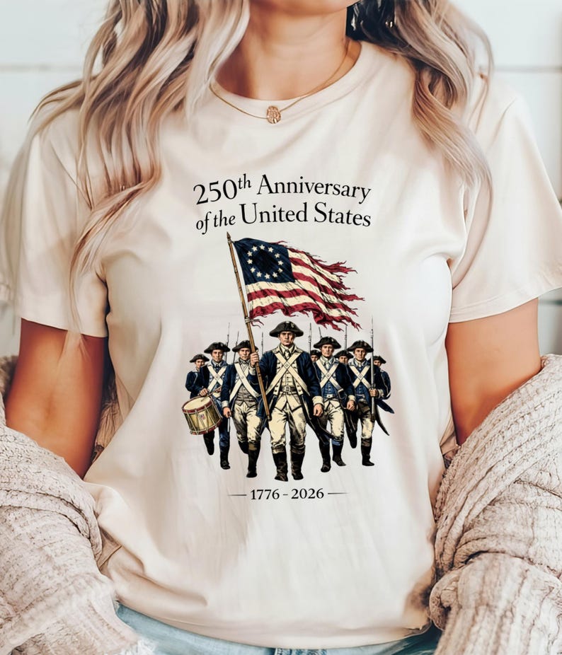 May include: Cream-colored t-shirt featuring a graphic commemorating the 250th Anniversary of the United States. The design includes an illustration of soldiers, a flag, and the years 1776-2026.