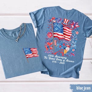 May include: Two blue jean t-shirts. One has a small American flag on the pocket, and a silver chain. The other has a large American flag design on the back with the text "250th Anniversary The United States of America 1776-2026".