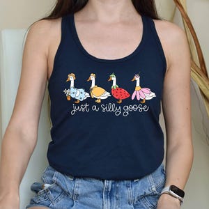 K&ouml;nnte beinhalten: Marineblaues Tanktop mit einer skurrilen Illustration von vier G&auml;nsen in verschiedenen Outfits, darunter ein Blumenkleid, ein gelber Mantel, ein Erdbeerkost&uuml;m und ein rosa Rock. Der Text "just a silly goose" steht darunter.