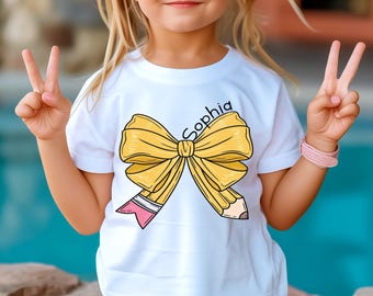 May include: White t-shirt featuring a large yellow bow and pencil design. The word "Sophia" is written in cursive above the bow. The pencil has a pink eraser. A child is wearing the shirt and giving the peace sign.