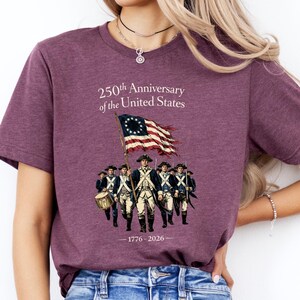 May include: A maroon t-shirt featuring a graphic commemorating the 250th Anniversary of the United States. The design includes a waving American flag, a group of soldiers, and the years 1776-2026.