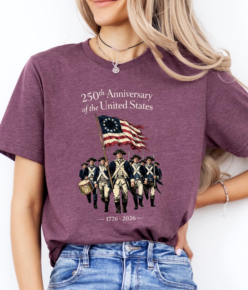 May include: A maroon t-shirt featuring a graphic commemorating the 250th Anniversary of the United States. The design includes a waving American flag, a group of soldiers, and the years 1776-2026.
