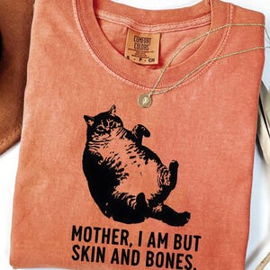 May include: A yam-colored t-shirt with a black graphic of a cat lying on its back. The text "MOTHER, I AM BUT SKIN AND BONES" is printed below the cat. A gold necklace with a pendant is draped across the shirt.
