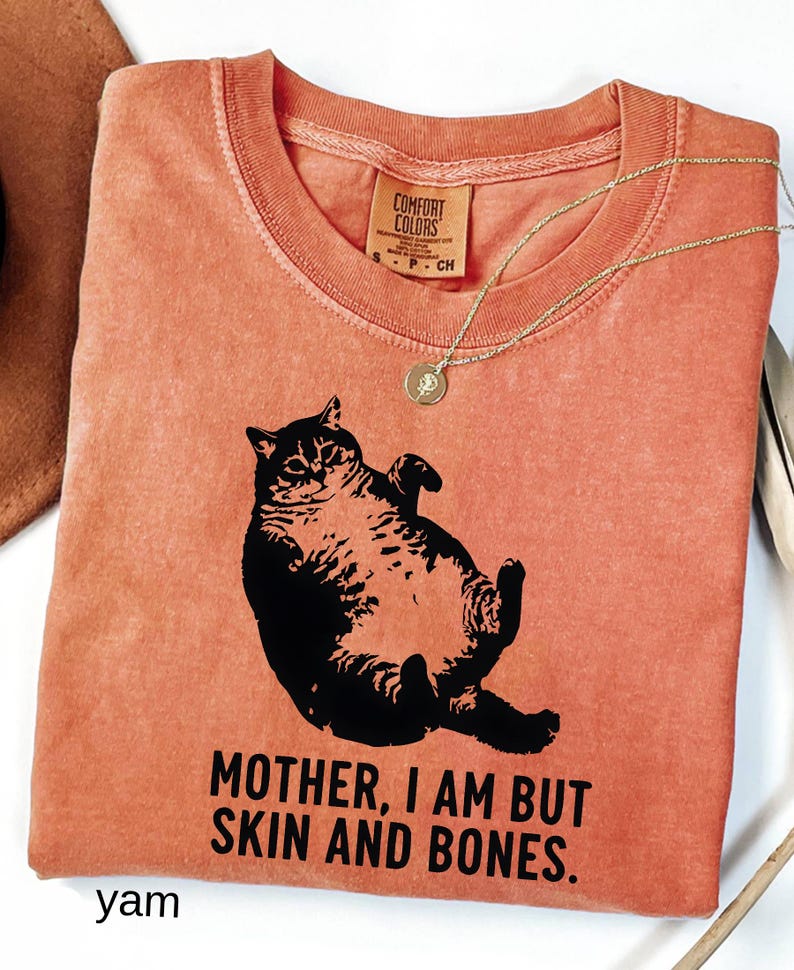 May include: A yam-colored t-shirt with a black graphic of a cat lying on its back. The text "MOTHER, I AM BUT SKIN AND BONES" is printed below the cat. A gold necklace with a pendant is draped across the shirt.