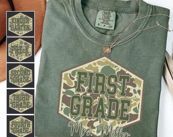 Può includere: T-shirt verde muschio con un design esagonale mimetico che recita "FIRST GRADE Mrs. Miller". Una collana con un ciondolo è sulla maglietta. Ulteriori disegni esagonali con livelli di classe sono a sinistra.