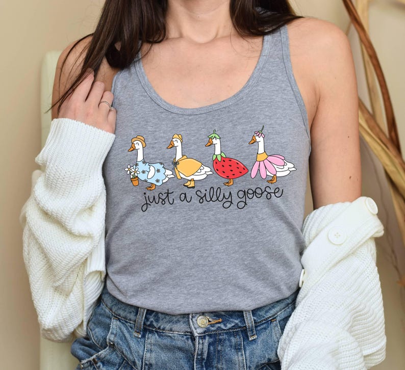 K&ouml;nnte beinhalten: Graues Tanktop mit einer skurrilen Illustration von vier G&auml;nsen in verschiedenen Outfits, darunter ein Blumenkleid, ein gelber Mantel, ein Erdbeerkost&uuml;m und ein rosa Rock. Der Text "just a silly goose" steht darunter.