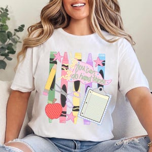 May include: A white short-sleeve t-shirt featuring a colorful graphic of school supplies including crayons, a pencil, scissors, an apple, and a clipboard. The design includes a pink banner with the text "you can do hard things" in green.