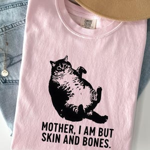 May include: A light pink t-shirt with a black graphic of a cat lying on its back. The text "MOTHER, I AM BUT SKIN AND BONES" is printed below the cat. The word "Blossom" is printed at the bottom.