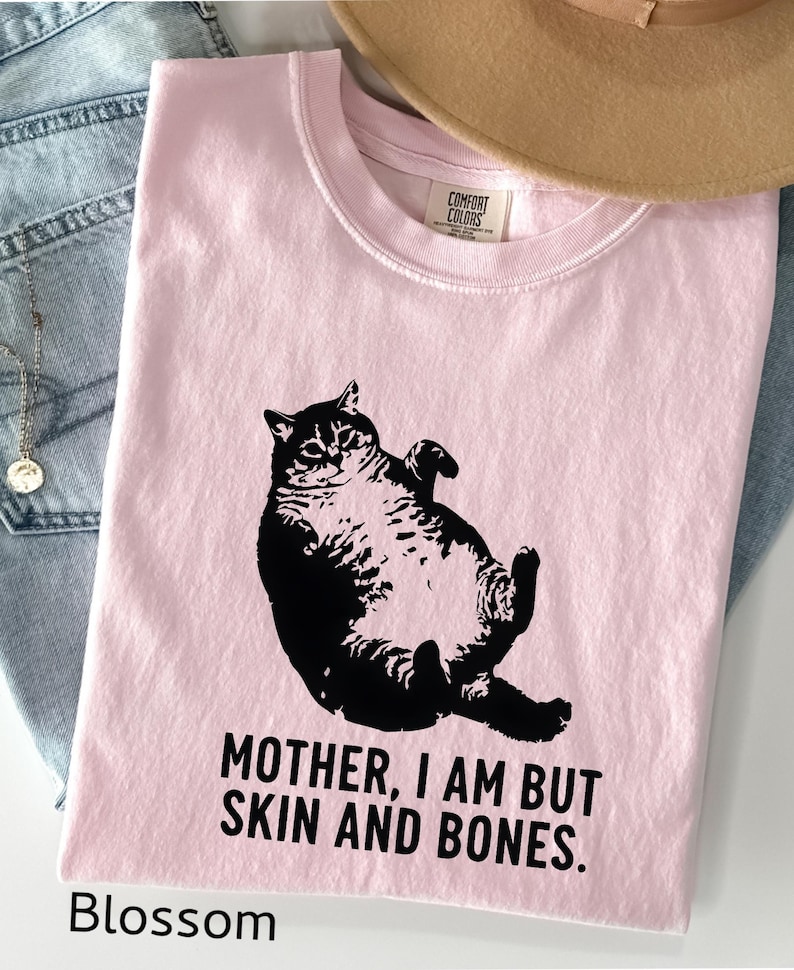 May include: A light pink t-shirt with a black graphic of a cat lying on its back. The text "MOTHER, I AM BUT SKIN AND BONES" is printed below the cat. The word "Blossom" is printed at the bottom.