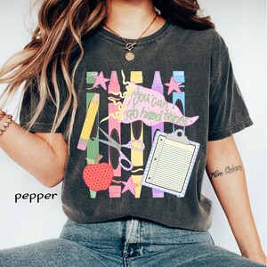 May include: A charcoal gray short-sleeve t-shirt featuring a graphic print of colorful crayons, a red apple, scissors, a clipboard, and a pink pennant flag with the text "you can do hard things" written in a cursive font with small stars.