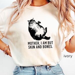 May include: Ivory-colored t-shirt featuring a black graphic of a cat lying on its back. The text "MOTHER, I AM BUT SKIN AND BONES" is printed below the cat. The word "ivory" is printed on the bottom right.
