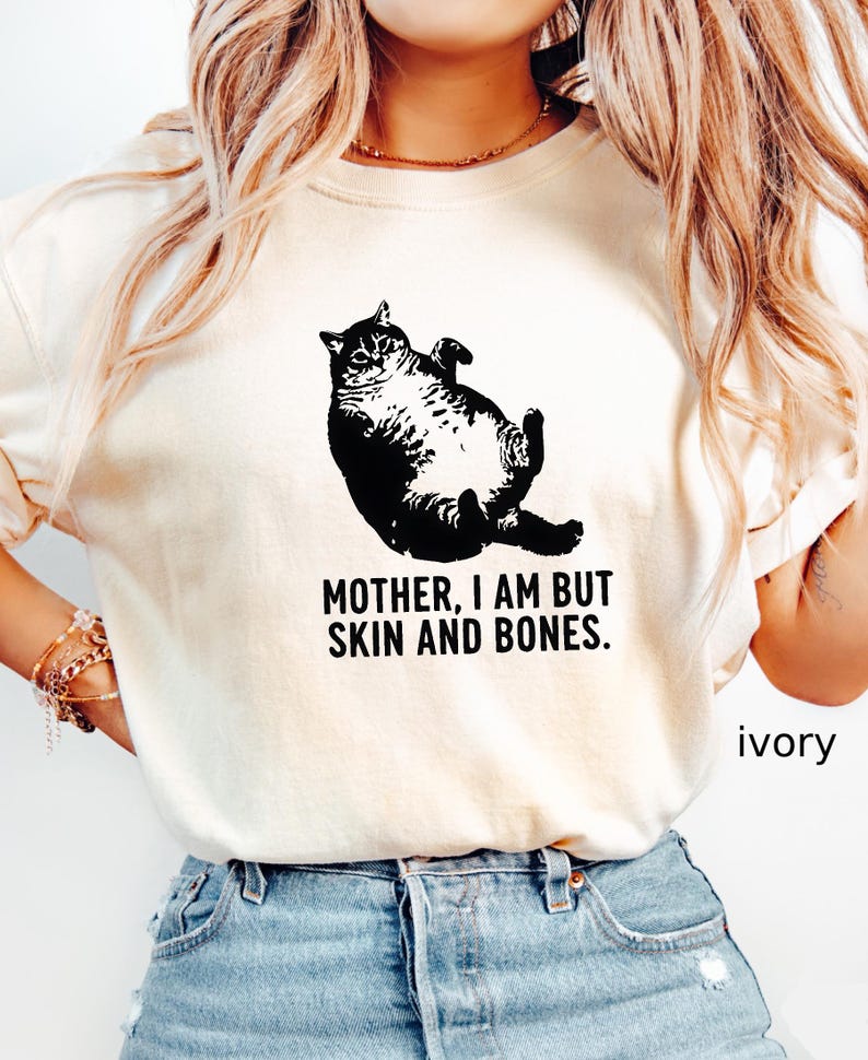 May include: Ivory-colored t-shirt featuring a black graphic of a cat lying on its back. The text "MOTHER, I AM BUT SKIN AND BONES" is printed below the cat. The word "ivory" is printed on the bottom right.