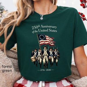May include: Forest green t-shirt with a graphic commemorating the 250th Anniversary of the United States. The design features a colonial flag and soldiers, with the years 1776-2026. The text "250th Anniversary of the United States" is printed above the graphic.