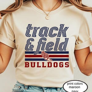 May include: Cream-colored t-shirt with the words "track & field" in navy blue, and "BULLDOGS" in maroon. A winged shoe graphic is featured. The print colors are maroon and navy.