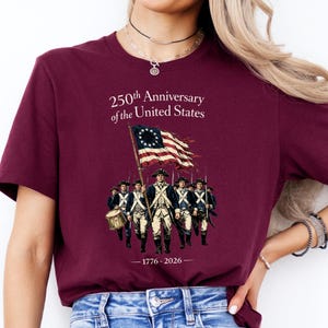 May include: Burgundy t-shirt featuring a graphic commemorating the 250th anniversary of the United States. The design includes an illustration of soldiers marching with a flag, and the years 1776-2026.