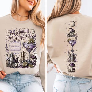 May include: A beige crewneck sweatshirt featuring a purple and black graphic print. The front displays the text Midnight Margaritas with a cocktail glass, a teapot, a frog in a witch hat, and mushrooms. The back shows a vertical arrangement of these icons.