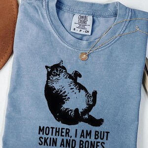 May include: A blue jean Comfort Colors t-shirt featuring a black graphic of a cat lying on its back. The text "MOTHER, I AM BUT SKIN AND BONES" is printed below the cat. A gold necklace with a pendant is draped on the shirt.