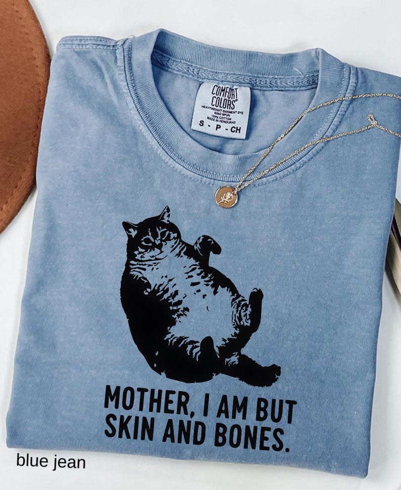 May include: A blue jean Comfort Colors t-shirt featuring a black graphic of a cat lying on its back. The text "MOTHER, I AM BUT SKIN AND BONES" is printed below the cat. A gold necklace with a pendant is draped on the shirt.